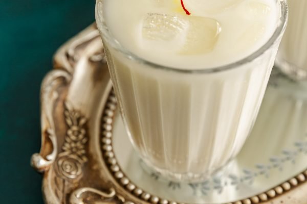 Glas mit kalter Safran-Kardamom-Horchata, verziert mit Zimtstange und goldenen Safranfäden auf Eisklötzen.